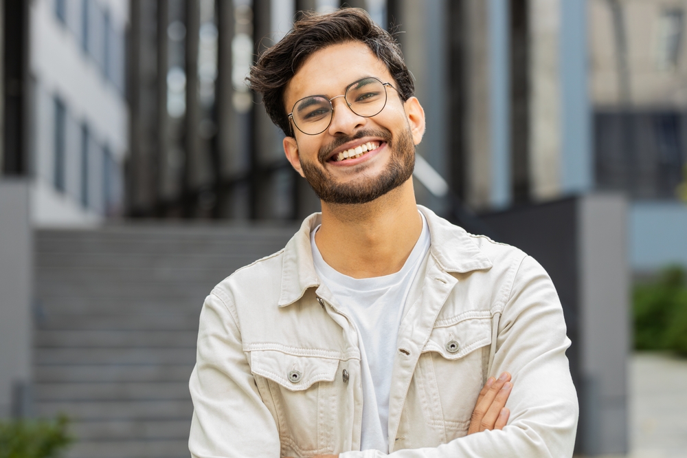 Portrait of happy young Indian man smiling friendly, glad expression looking at camera dreaming, resting relaxation feel satisfied good news outdoors. Arabian Hindu guy in city street. Business people